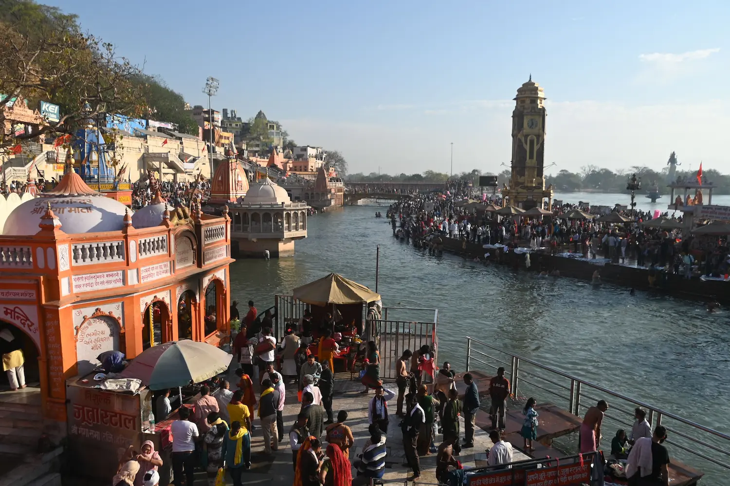 Panoramic daytime view of Har Ki Pauri ghat in Haridwar showing the iconic clock tower, orange temples, and devotees along the River Ganges.